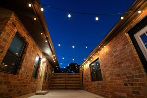 A brick building with a covered outdoor area at dusk, string lights overhead, and a wooden gate. - Canyon Lake · 4BR Home + Suite · Sleeps 10 · Pets OK (Canyon Lake)