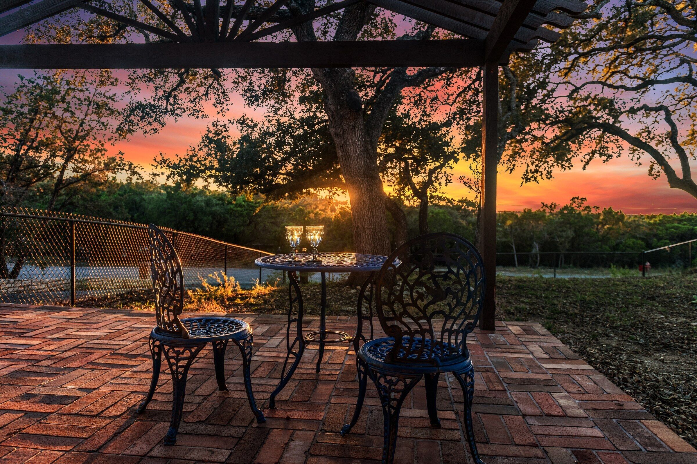 An outdoor patio with a pergola, two metal chairs, a table, and a view of a sunset.