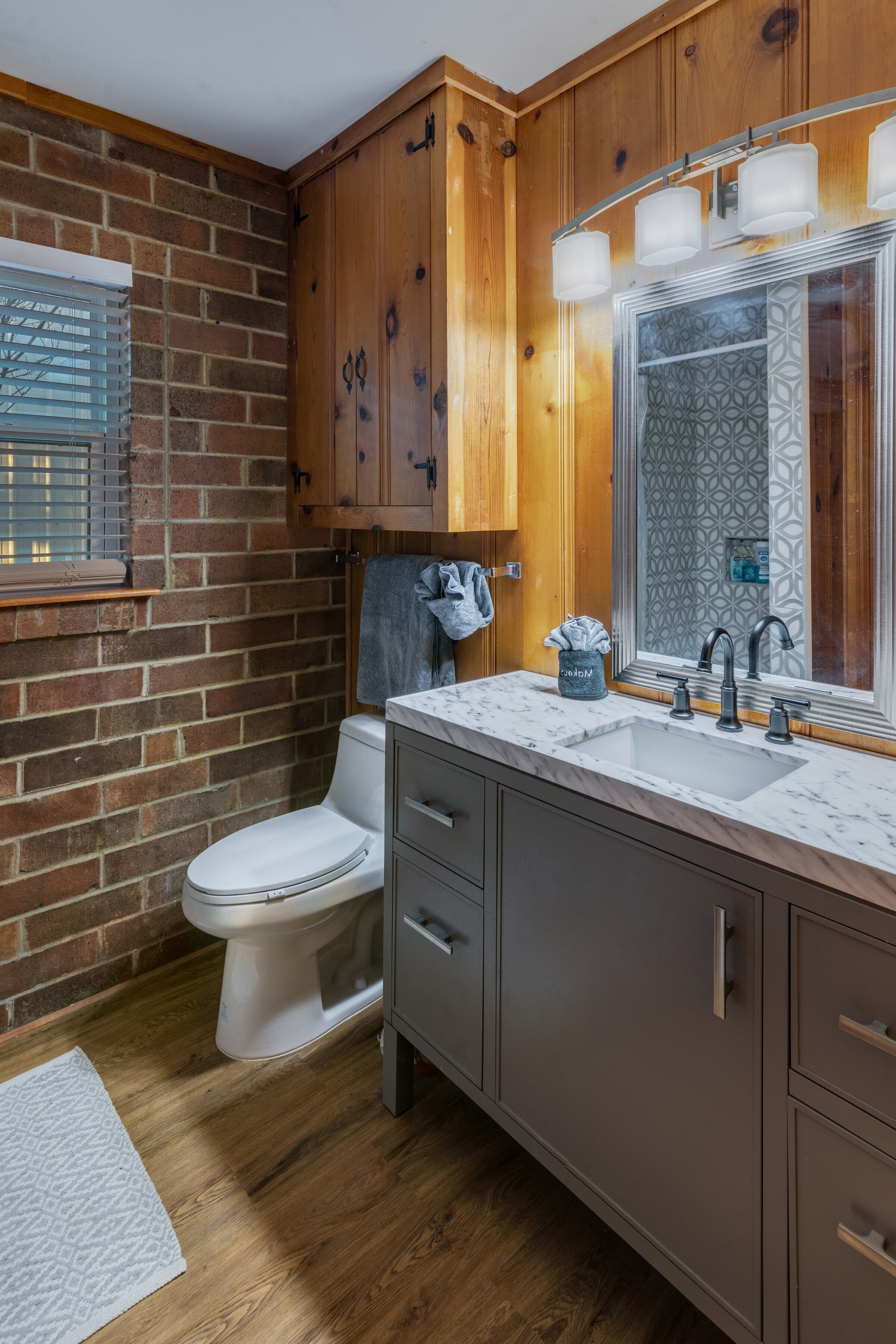 A bathroom with a toilet, a sink with a mirror, wooden cabinets, and a window with blinds.