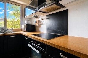 A modern kitchen with a black stove, stainless steel range hood, and a window with a view of greenery. - Appartement Sérénité Charonne - Welkeys (Paris)
