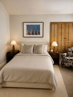 Een slaapkamer met een groot bed, twee nachtkastjes met lampen, een stoel, een raam met luiken en een lijst met een afbeelding aan de muur.