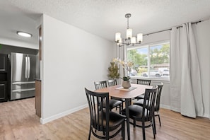 A modern kitchen with stainless steel appliances, a dining area with a wooden table and black chairs, and a window with curtains.