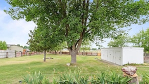 Ein Garten mit einem großen Baum, einer Holzterrasse und einem Gerätehaus.