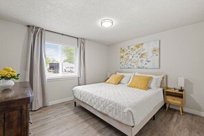 A bedroom with a bed, a nightstand, a lamp, a window with curtains, and a floral painting on the wall.