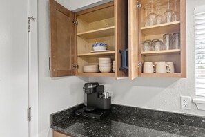 Een keuken met een koffiezetapparaat op het aanrecht, een kastje met servies en glazen, en een raam met jaloezieën.