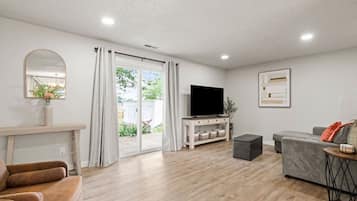 Una sala de estar moderna con televisor de pantalla plana, sofá gris, piso de madera y puerta corrediza de vidrio que da a un espacio al aire libre.