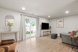 A modern living room with a flat-screen TV, grey sofa, wooden flooring, and a sliding glass door leading to an outdoor area.