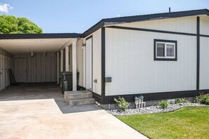 A single-story mobile home with a covered driveway, a small front porch, and a visible trash bin.