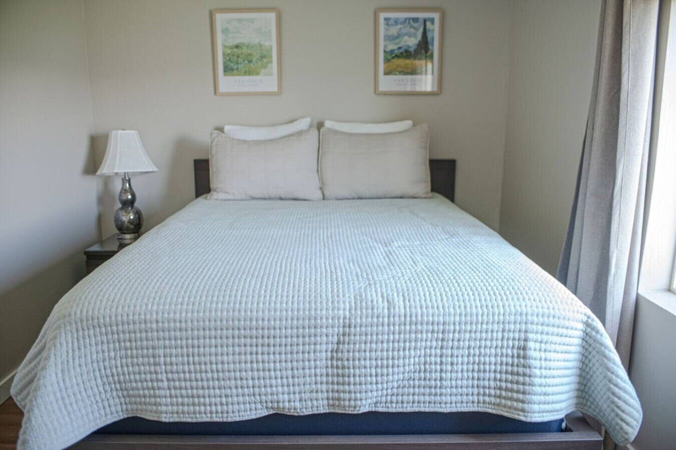 A standard hotel room with a bed, a nightstand with a lamp, and two framed pictures on the wall.
