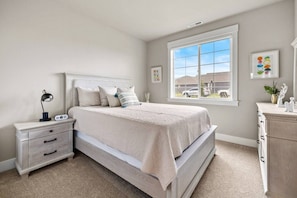 A bedroom with a bed, bedside table, lamp, and a window with a view of houses.