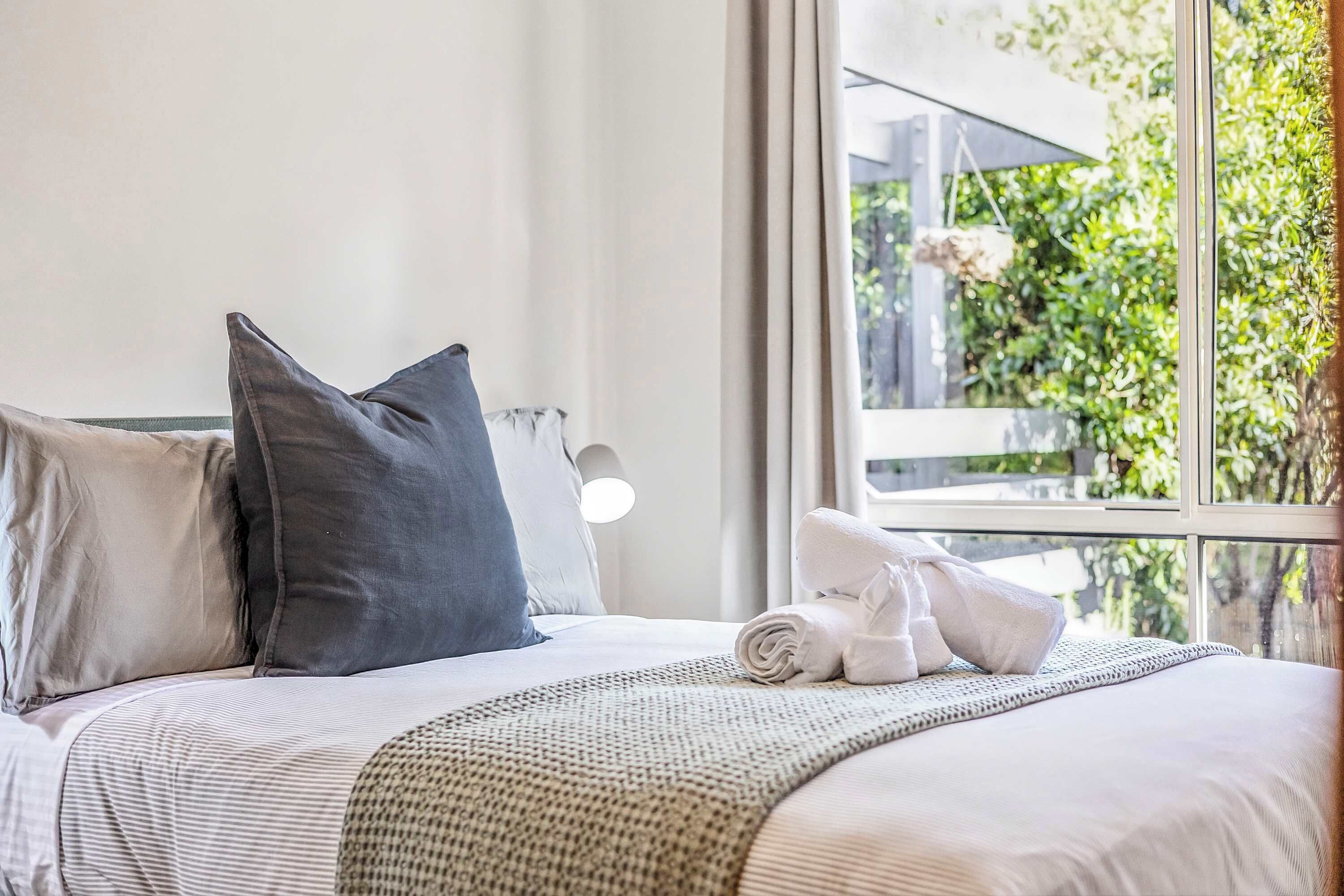 A neatly made bed with pillows and a rolled towel, a bedside lamp, and a window with a view of greenery.