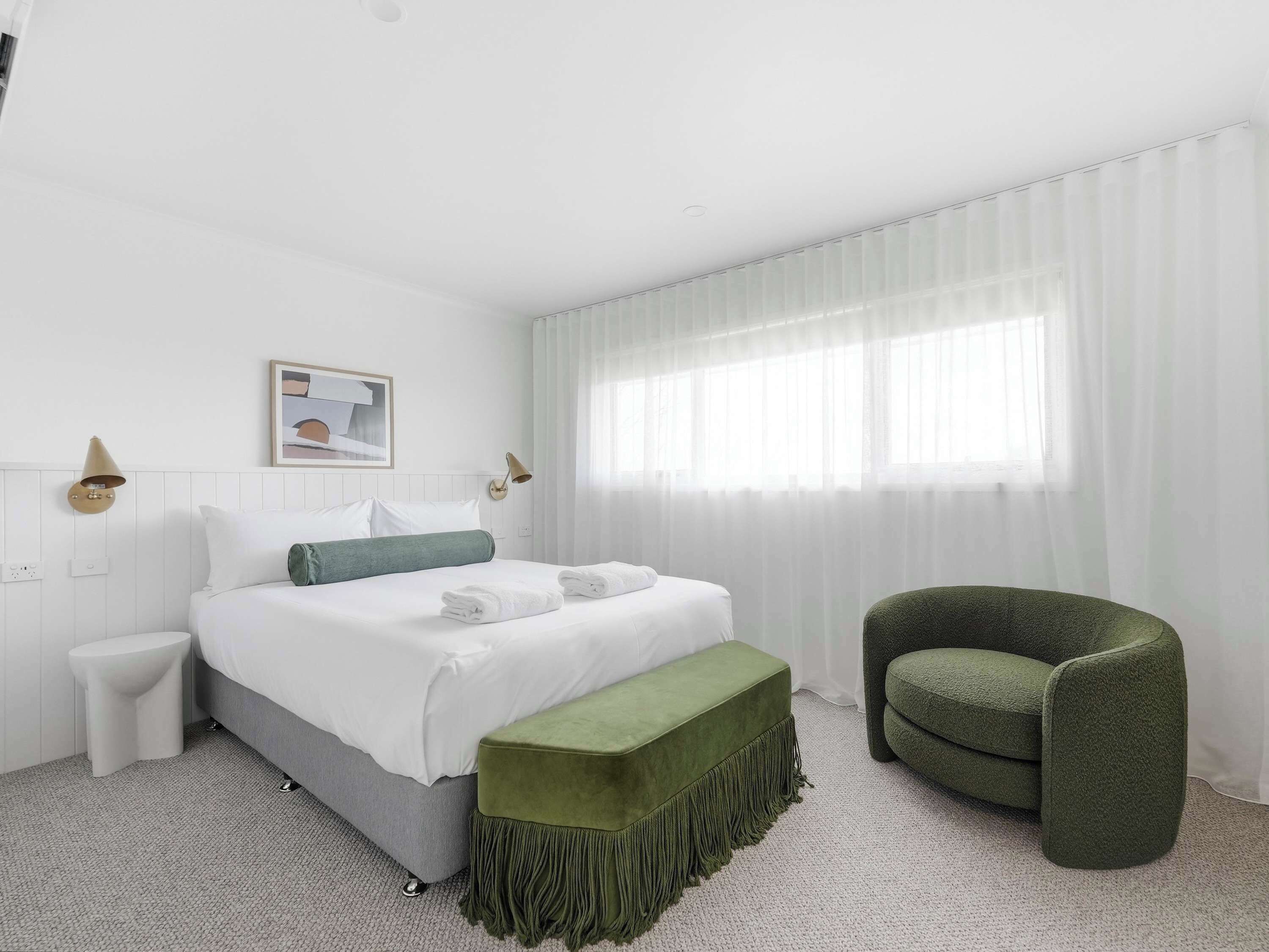 A hotel room with a bed, a green armchair, a small table, and a window with curtains.