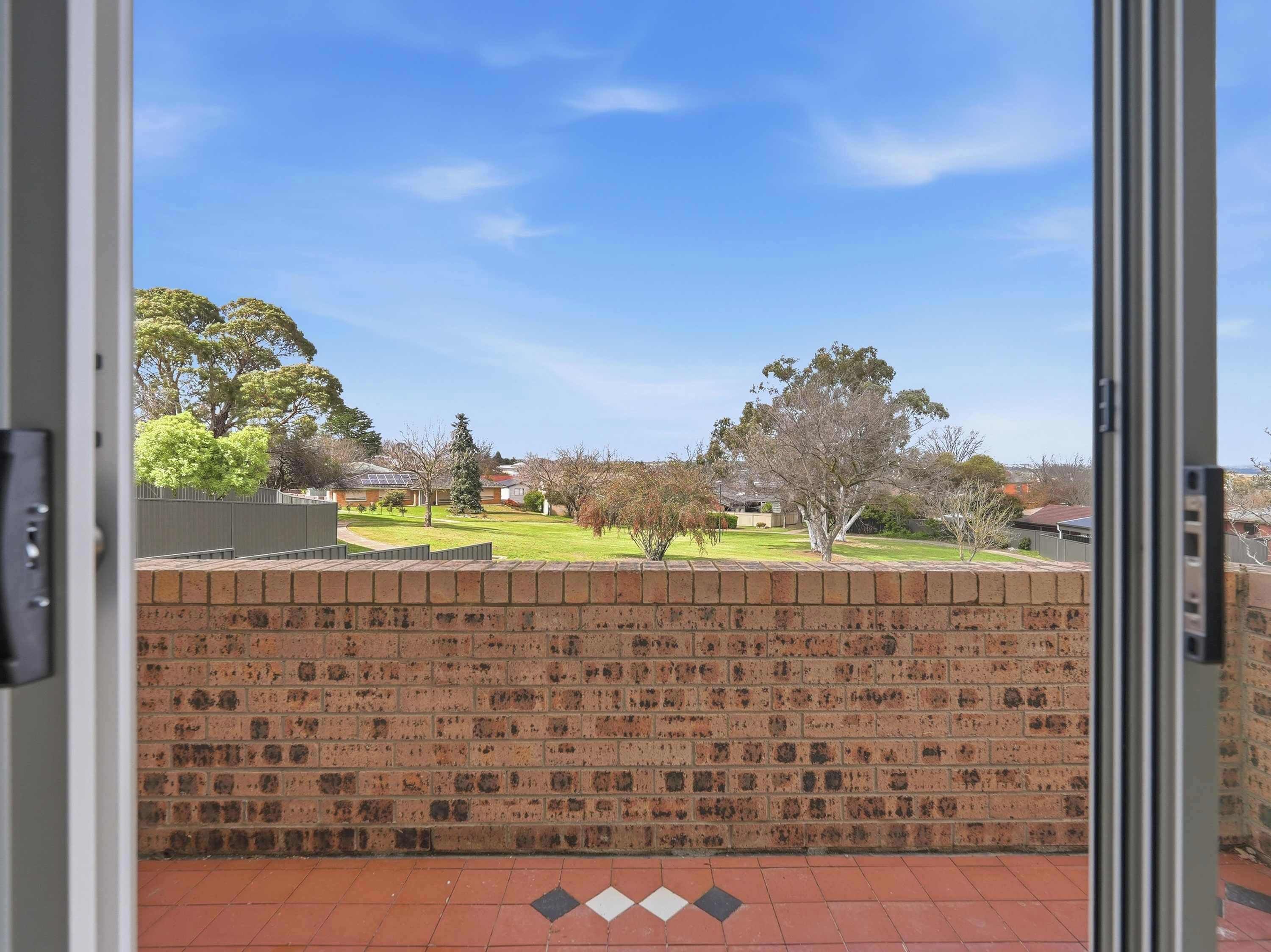 A view through a glass door of a brick wall with a tiled floor, leading to a grassy area with trees and houses in the background.