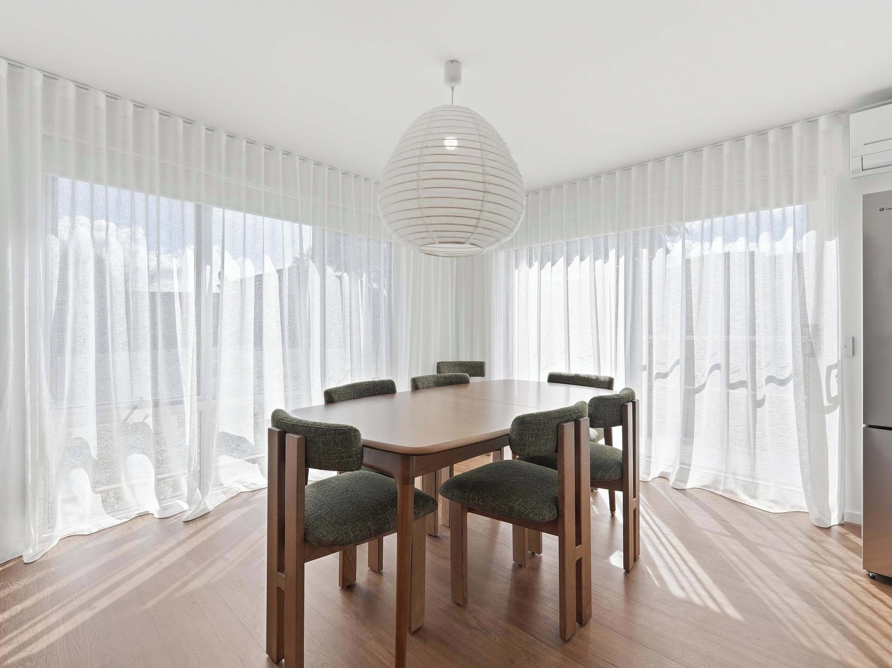 Un coin repas avec une table ronde et six chaises, de grandes fenêtres avec des rideaux légers et un luminaire suspendu moderne.