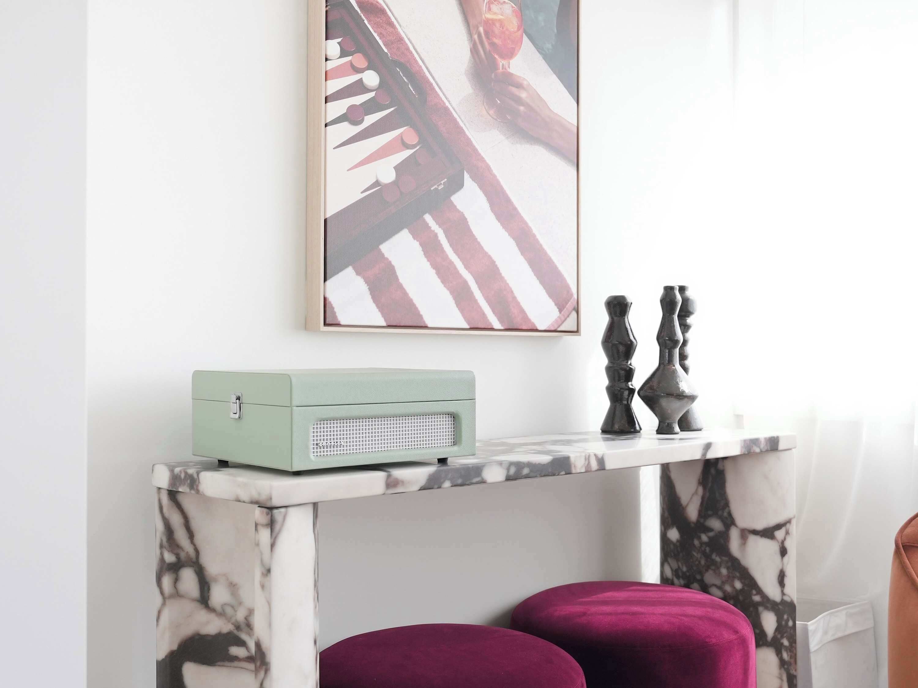 Une pièce comprenant une console dont le plateau est décoré d’un motif imitant le marbre, une radio ancienne et un tableau abstrait encadré.