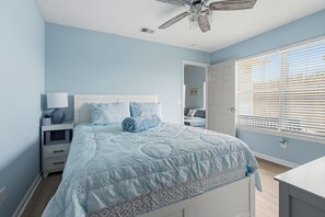 Un dormitorio con cama, mesita de noche, lámpara, ventilador de techo y ventana con persianas.