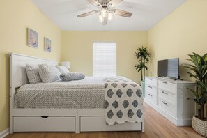A bedroom with a bed, a ceiling fan, a window with blinds, a TV on a dresser, and a potted plant. - So Cute! WALK to Beach Close to Restaurants Marina & State Park Fenced 4 Ur Dog! (Panama City Beach)