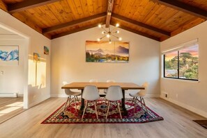 Een eetruimte met een houten tafel, zes stoelen, een kamerkleed met een patroon en een schilderij van een landschap.