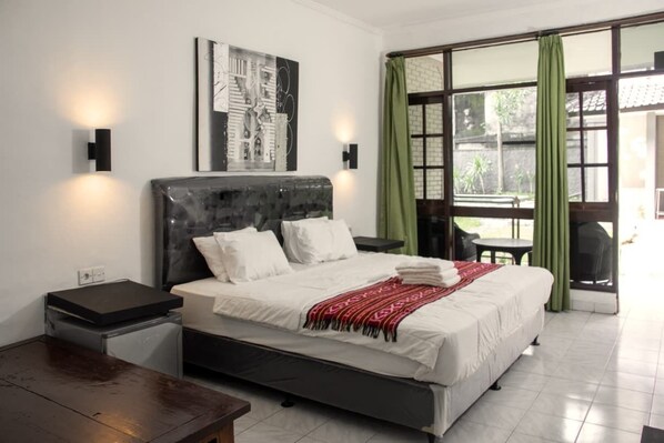 A hotel room with a bed, a nightstand, a small table, a window with curtains, and a wall-mounted artwork. - Moanna Stay Kuta Beach (Kuta)