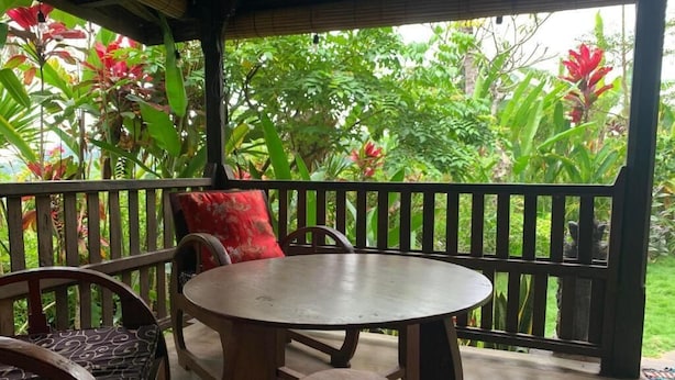 Una terraza de madera con una mesa redonda y dos sillas, rodeada por una barandilla negra, con vista a un jardín exuberante con diversas plantas y flores.