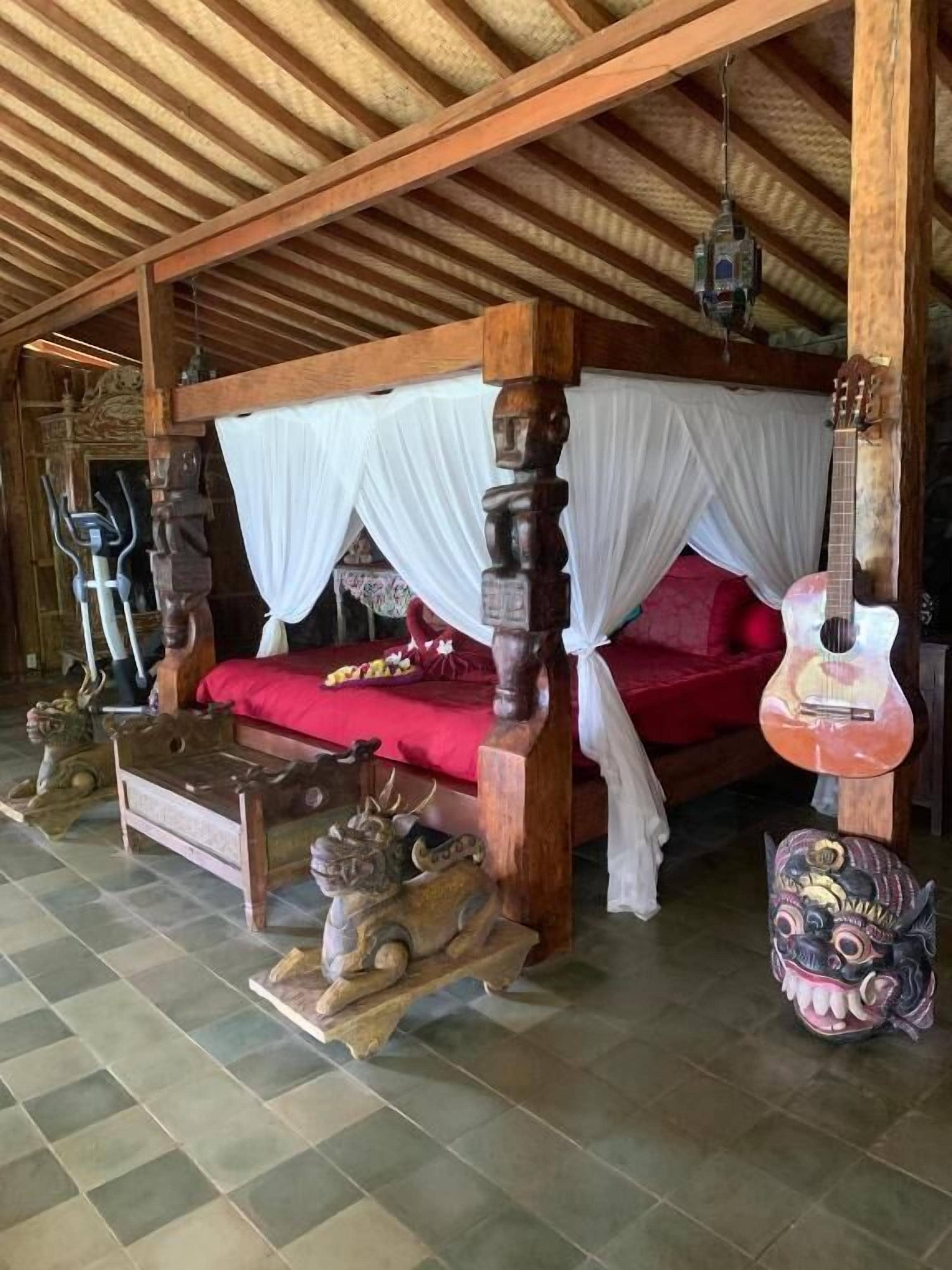 A four-poster bed with a red bedspread, a guitar, and decorative items on the floor.