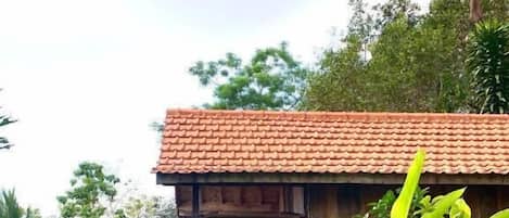 A wooden house with a tiled roof, surrounded by lush greenery and a blue fence.