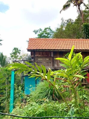 A wooden house with a tiled roof, surrounded by lush greenery and a blue fence. - villa kepuh (buleleng)