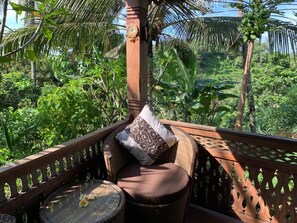 Una cadira de vimet amb un coixí i una taula petita rodona, col·locada en un balcó amb vista a vegetació tropical.