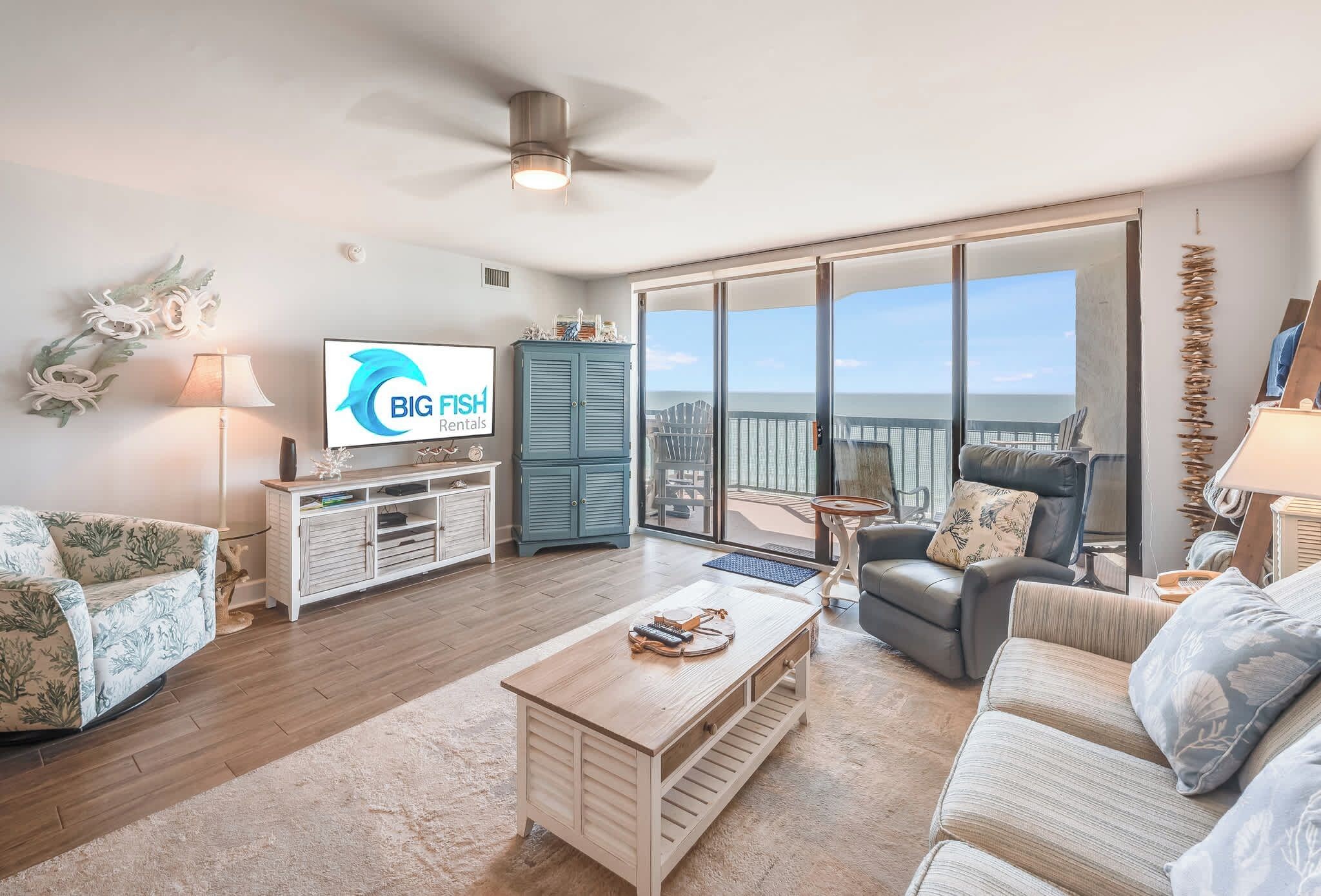 A spacious living room with a large sliding glass door leading to a balcony, a flat-screen TV mounted on the wall, and a ceiling fan.