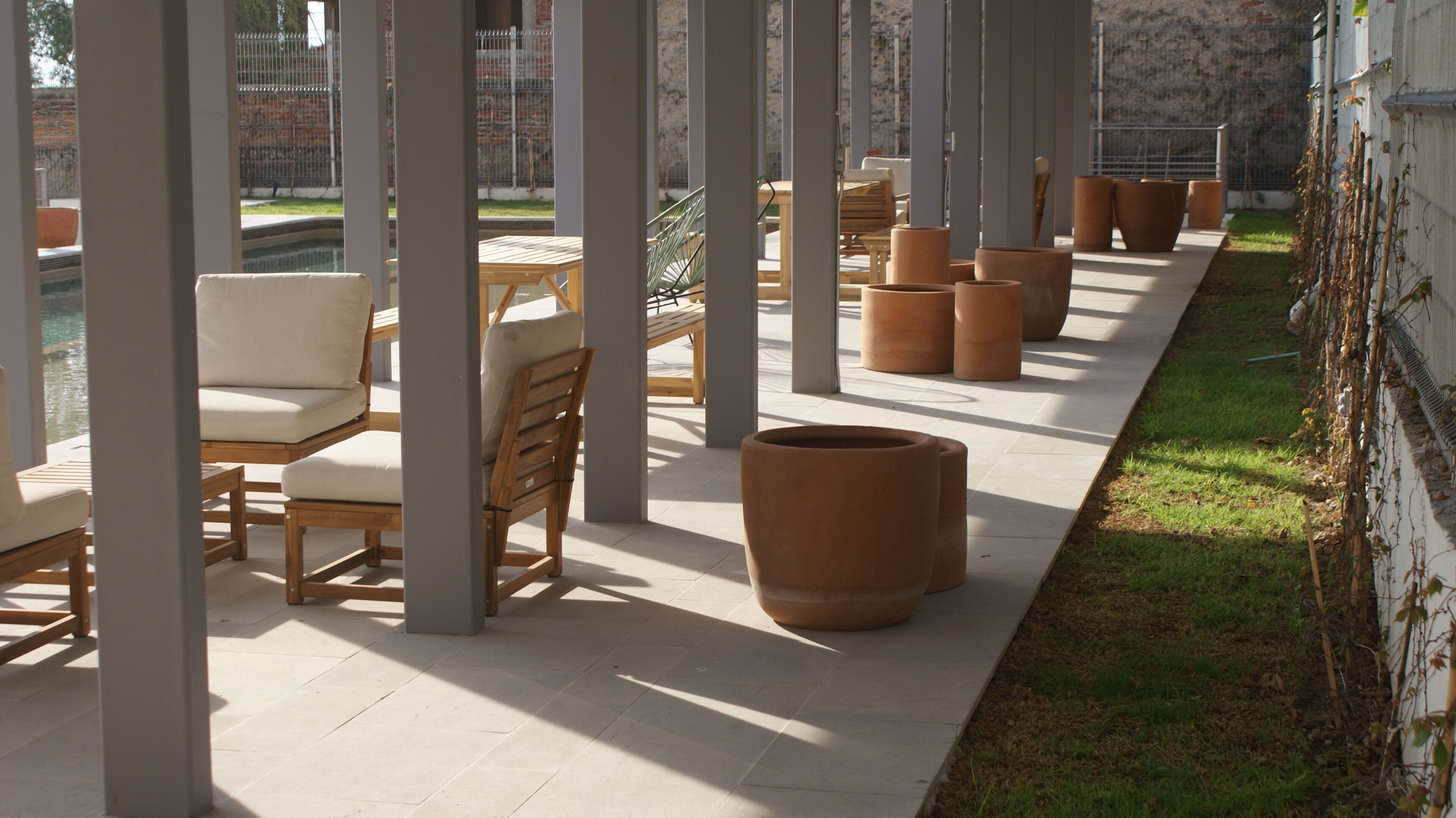 Un espacio al aire libre cubierto con sillas, macetas grandes, piso de baldosas y vistas a una piscina y jardín.