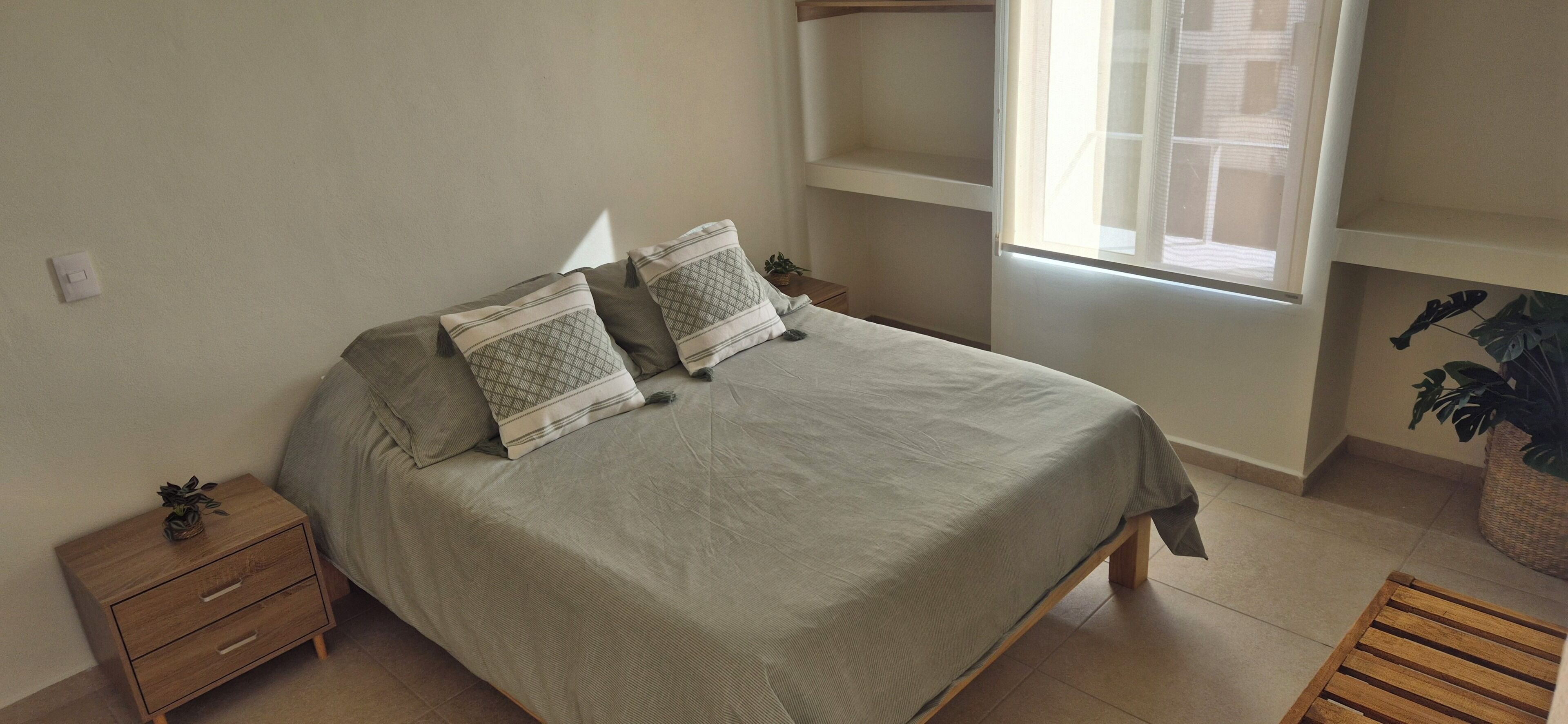 Un dormitorio con una cama, dos mesitas de noche, un banco de madera, una ventana y una planta.