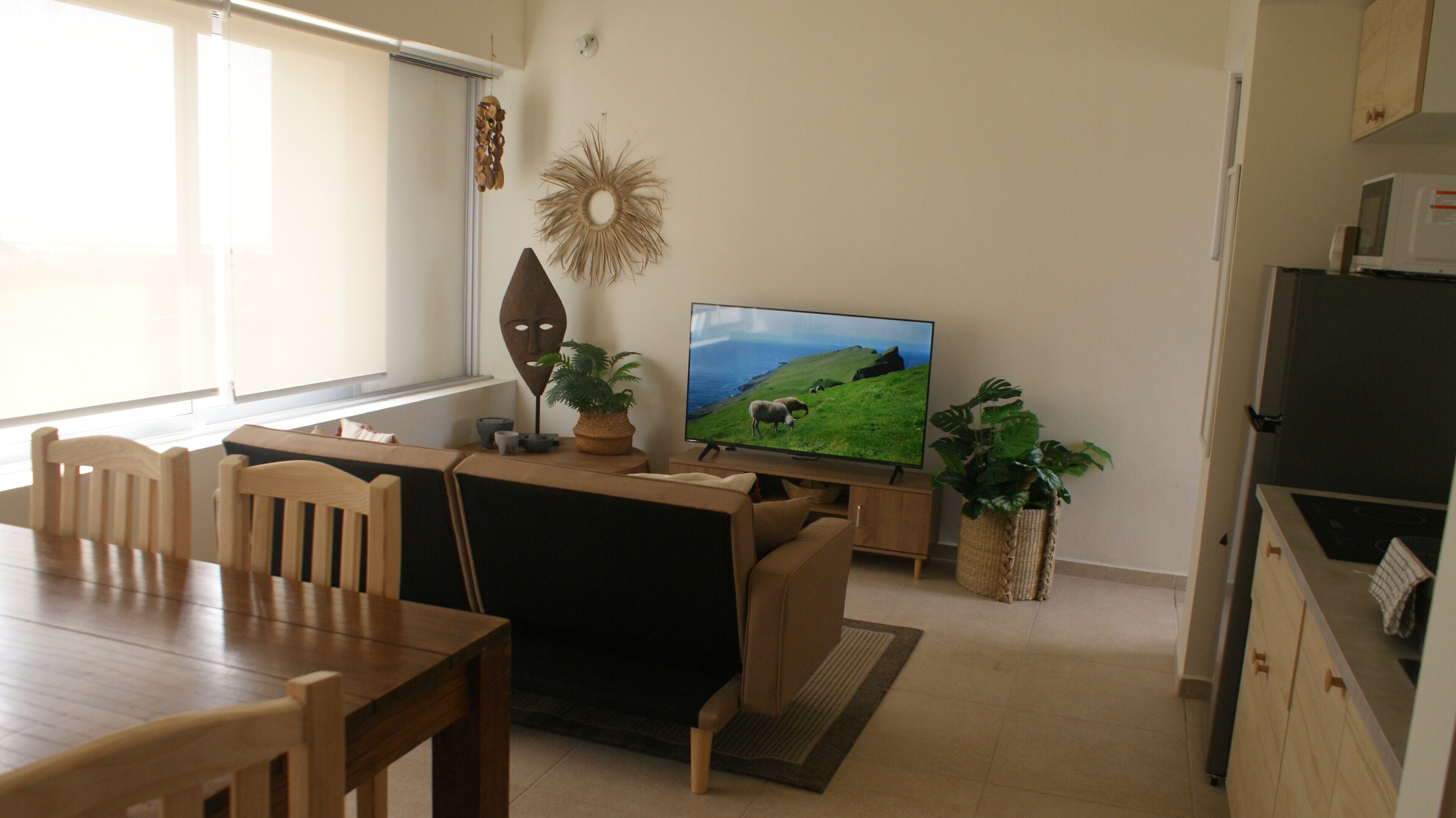 A living room with a wooden dining table, a sofa, a television displaying a nature scene, and a kitchen area with cabinets and a microwave.