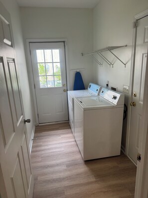 A laundry room with a front-loading washer and dryer, a door with a window, and a metal drying rack on the wall. - Your Home Away From Home Under 10 Min to Ft. Benning & 15 min to Uptown Columbus (Columbus)