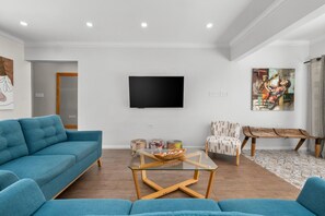 A living room with a blue sofa, a glass coffee table, a TV mounted on the wall, and a patterned chair.