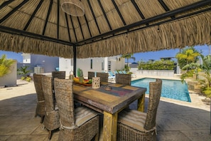 An outdoor dining area with wicker furniture and a thatched roof, overlooking a swimming pool and a modern building. - Private 3BR2BA Villa w/ Private Pool & Close2Beach (Noord)