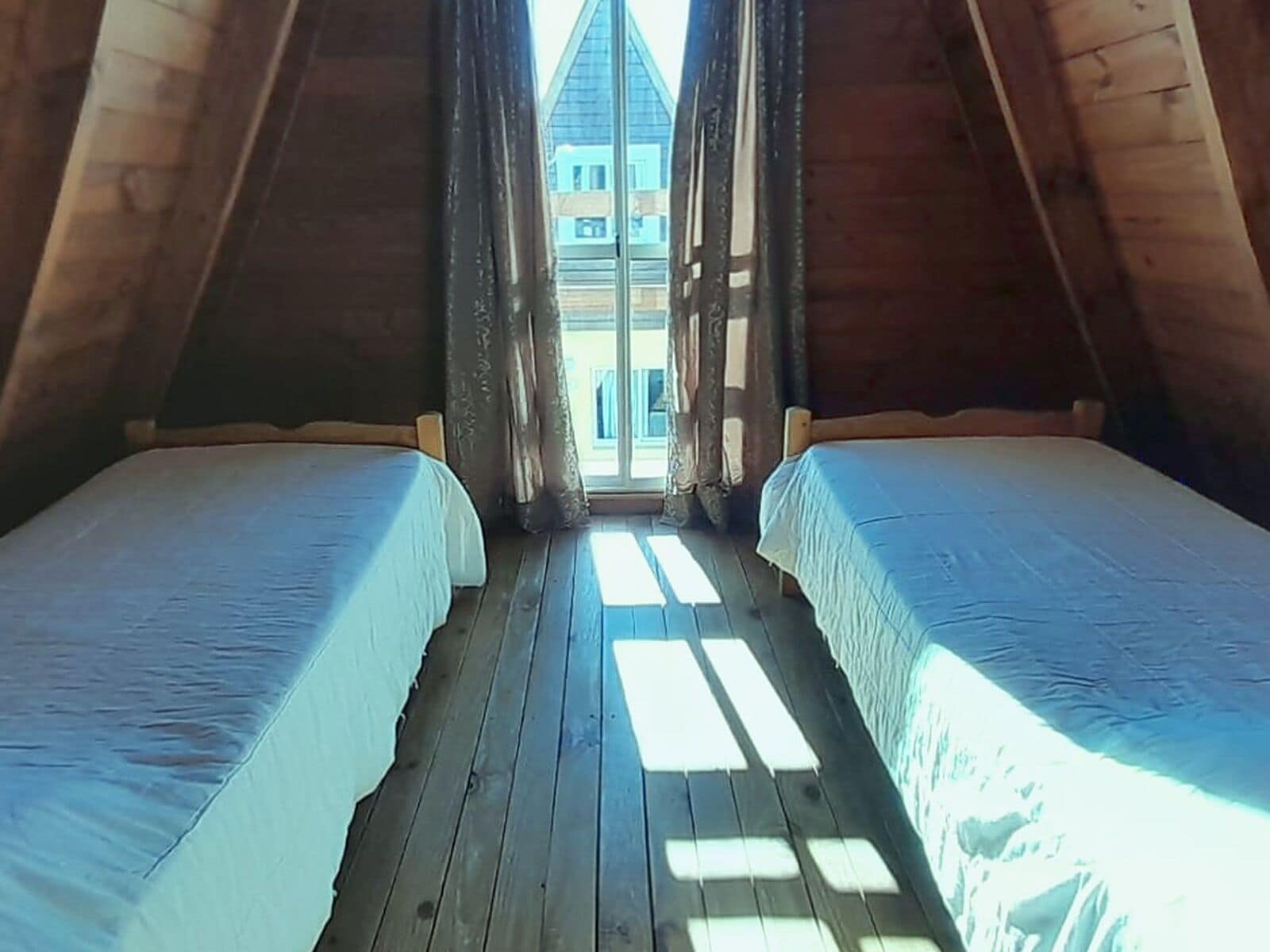 Quarto com duas camas alinhadas, piso de madeira e vista para um edifício através da janela.