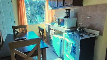 Una cocina compacta con estufa a gas, microondas y lavamanos. Hay una mesa de comedor con sillas y una ventana con cortinas.