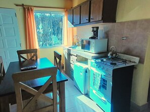 A compact kitchen with a gas stove, microwave, and a sink. There is a dining table with chairs and a window with curtains.