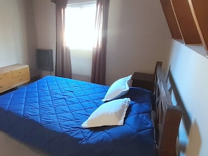 A bedroom with a bed, a wooden dresser, a nightstand, and a window with curtains.