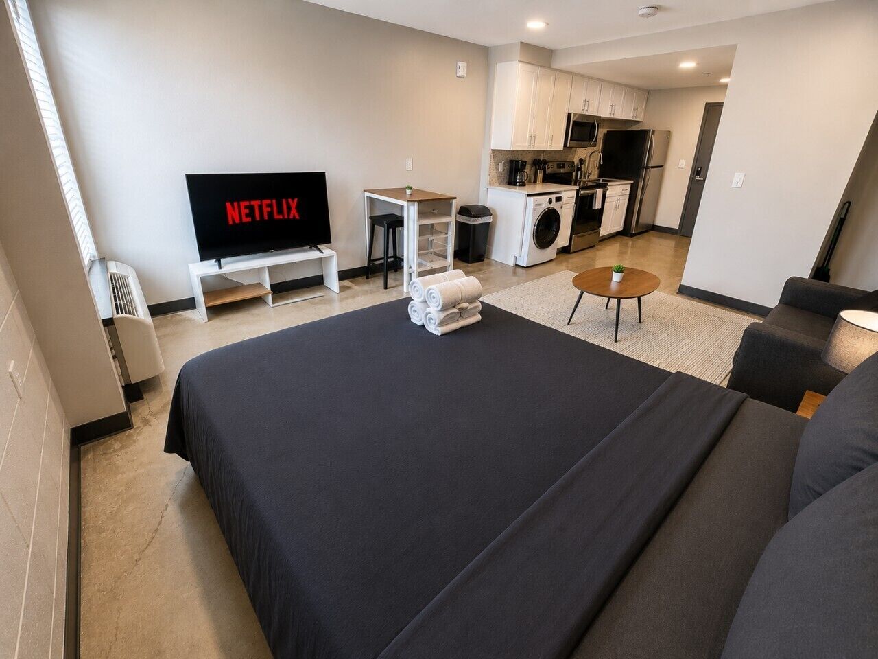 A modern hotel room with a large bed, a TV, a small kitchen area, and a seating area.
