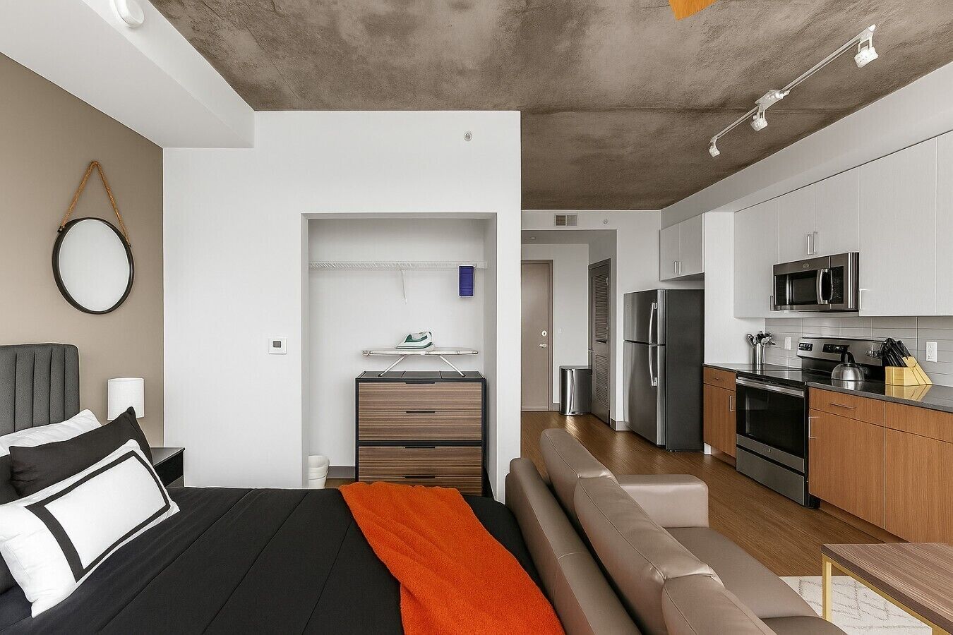 A modern hotel room with a bed, a seating area, a kitchenette, and a bathroom.