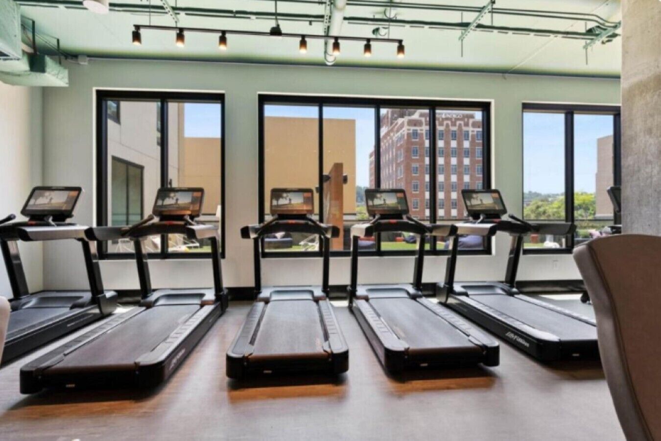 A gym with treadmills aligned in rows, large windows, and a view of buildings outside.