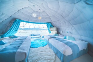 Una habitación con techo abovedado, dos camas, un tapete blanco y un ventanal con cortinas.