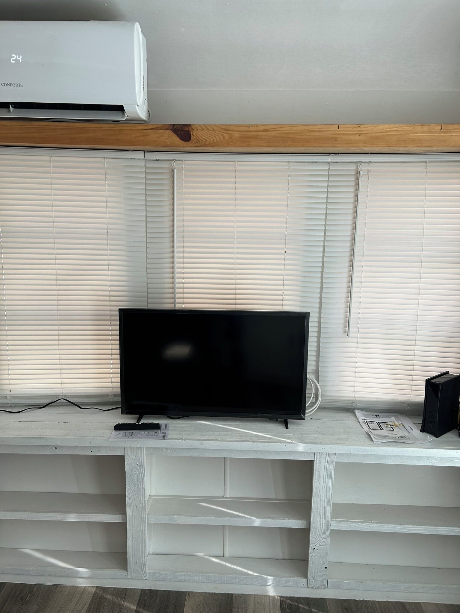 Un televisor de pantalla plana sobre un estante blanco, un aire acondicionado blanco arriba y persianas blancas en las ventanas.