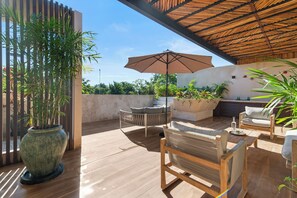 Une terrasse extérieure avec un plancher en bois, une grande plante en pot, un espace salon circulaire avec des coussins et une table en bois surmontée d’un parasol.