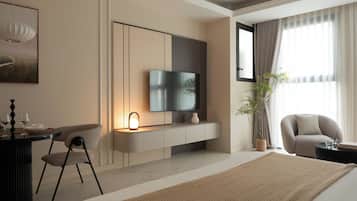 A modern hotel room with a flat-screen TV, a dining area with a table and chairs, a large bed, and a potted plant.