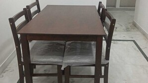 Una mesa rectangular con cuatro sillas, una pared blanca y una zona de cocina con estantes.