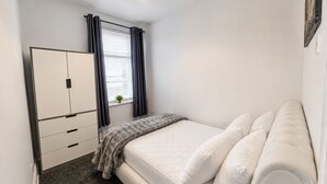 Ein Schlafzimmer mit Bett, Kleiderschrank, Fenster mit Vorhängen und einer kleinen Pflanze auf dem Fensterbrett.