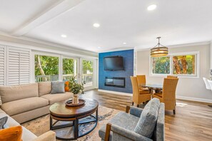 Ein geräumiges Wohnzimmer mit einer Couch, Sesseln, einem Couchtisch und einem Essbereich mit Korbstühlen. Ein Flachbildfernseher ist an der Wand befestigt, und große Fenster bieten Ausblicke ins Freie.