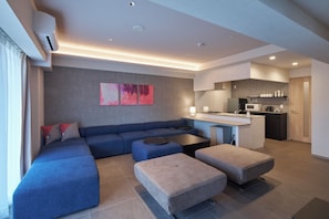 A modern living room with a sectional sofa, ottomans, a coffee table, and a kitchen area with a microwave and coffee machine. - KOUSA Osaka Namba (Osaka)